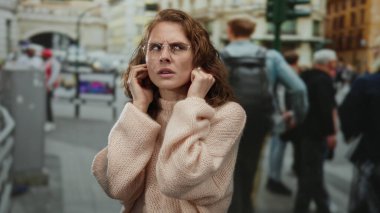 Şehir sokaklarında kulaklarını tıkayan gözlüklü ve süveterli bir kadın, kentsel arka planla çevrili ve insanlar gürültü dolu bir ortamda yürüyorlar, endişe ve rahatsızlıklarını dile getiriyorlar..