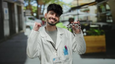 Beyaz önlüklü genç adam, canlı bir terasta gülümserken elinde optik aletle tıbbi ya da bilimsel bir bağlam öneriyor..