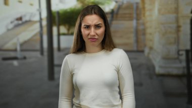 Beyaz tişörtlü genç sarışın kadın şehir sokaklarında, şehir mimarisinin ve yeşilliğin ortasında gün ışığında düşünceli bir ifadeyle dikiliyor..