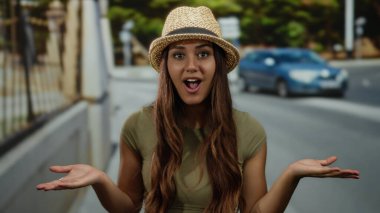 Şapkalı genç bir İspanyol kadın, elleri yüzü üzerinde bir caddede duruyor ve canlı bir şehir ortamında sürpriz yapıyor..