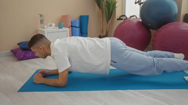 Spor salonunda egzersiz yapan genç bir adam egzersiz sırasında spor salonunda spor topları ve ekipmanlarla mavi minder üzerinde ahşap yapıyor.