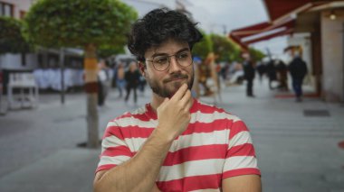 Çizgili gömlekli, sakallı ve gözlüklü genç adam bulanık arka planı ve ağaçları olan yoğun bir sokakta dalgın dalgın duruyor..