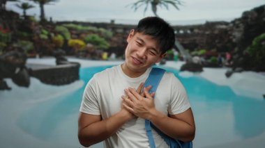 Sırt çantalı genç adam lüks havuzda sıcacık gülümsüyor. Bölgeyi çevreleyen tropikal bitkilerle dolu tesiste sakin bir atmosfer yaratıyor.
