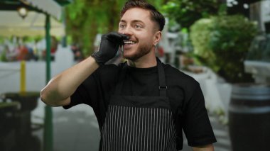 Sakallı ve önlüklü genç beyaz adam, bir şehir caddesinde garsonluk yapıyor, açık şehir ortamında eldivenle gülümsüyor..