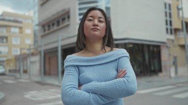 Mavi kazaklı kadın şehir binaları ve bulutlu gökyüzü ile şehir caddesinde güven içinde kollarını kavuşturarak açık havada gençlik özgüvenini yakalıyor..