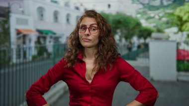 Kırmızı gömlekli kendine güvenen bir kadın şehir sokaklarında silah akimbo 'suyla dikiliyor, açık hava ortamında kararlılık ve kendine güvenini iletiyor..