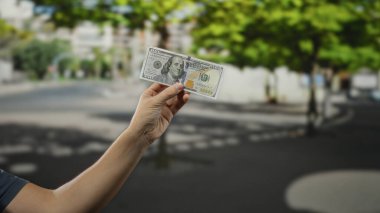Şehirdeki arka planda bulanık Amerikan doları banknotunu tutan adam, şehir ortamında ekonomik değişimi vurguluyor..