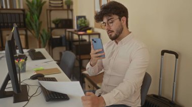 Ofisteki sakallı genç İspanyol adam bilgisayar ve bitkiler gibi modern çalışma alanlarıyla çevrili bir belgeyi ele geçirmek için akıllı telefon kullanıyor..