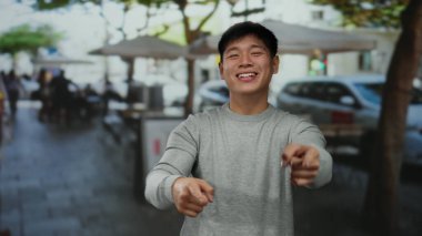 Genç Asyalı adam güneşli bir restoranın terasında otururken neşeli bir şekilde gülümsüyor ve arka planda insanlar var..