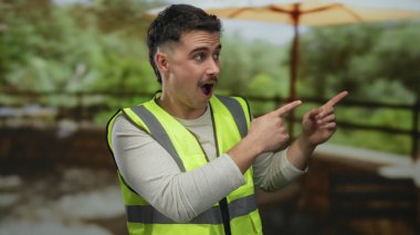 Yansıtıcı yelek giyen genç adam, geniş bir şemsiye altında yemyeşil bir park ortamında açık havada merak uyandırarak şaşırdı..