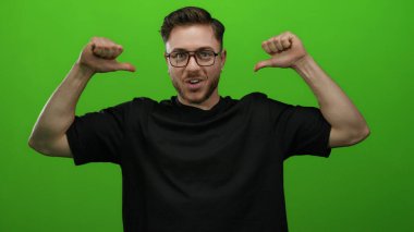 Gözlüklü ve sakallı beyaz bir adam, parlak yeşil bir arka planı olan, siyah tişörtlü ve kendinden emin bir şekilde gülümseyen birini işaret ediyor..