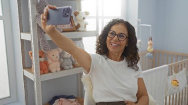 Yatak odasında içi doldurulmuş oyuncaklar ve beşiklerle dolu sıcak, neşe dolu iç mekan atmosferiyle selfie çeken bir kadın gülümsüyor..