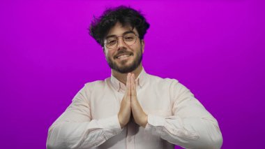 Gözlüklü ve sakallı genç adam mor arka planda gülümsüyor, açık renk gömlek giyiyor, pozitiflik ve sakin tavır takınıyor..