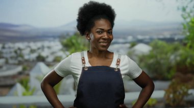 Önlüklü, arka planda şehir manzarası olan bir kadın güneşli bir günde şehir ortamında kendine güvenini sergiliyor..