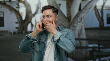 Kot ceketli genç adam parkta telefonla konuşuyor. Arka planda beyaz binalar ve ağaçlar bulanık. Mutluluğu ifade ediyor..