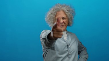 Gri saçlı kadın, mavi arka planda kameraya şakacı hareketler yaparak kendine güven ve mizah öneriyor..