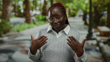 Gülümseyen ve el kol hareketi yapan bir kadın parkın içinde elleri ile davetkar ve neşeli bir hava sergiliyor.