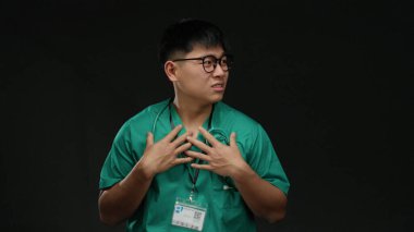 Siyah arka planda tek başına duran yeşil önlüklü genç adam profesyonel ortamda steteskop takıyor..