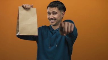 İzole edilmiş turuncu arka planda kameraya işaret eden İspanyol genç adam nişan ve etkileşim öneriyor.