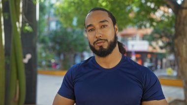 Sakallı genç adam şehir parkında duruyor, arka planda yeşil ağaçlar ve şehir binaları olan mavi bir gömlek giyiyor..