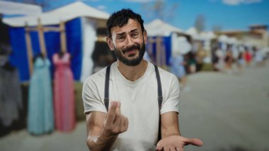 İspanyol sakallı adam sokakta jestler yapıyor. Arka planda bulanık piyasa var. Güneşli bir günde çadırlarla ve yakınlarda yürüyen insanlarla duygularını ifade ediyor..