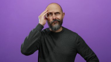 Gri kazaklı sakallı kel bir adam, alnında bir elle, izole edilmiş mor bir arka planda kafası karışmış gibi görünüyor..