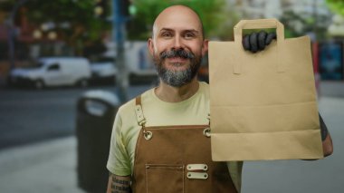 Kentsel caddede elinde kağıt torba olan, kahverengi tulumlu, açık havada gülümseyen, şehrin arka planında sergi açan bir adam..