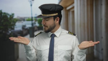 Pilot üniformalı, sakallı genç adam sorgu sırasında kollarını uzatarak dışarıda duruyor. Bulanık bir sokak geçmişi var ve yüzbaşı şapkası takıyor..