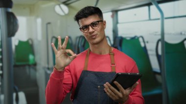 Kırmızı kazaklı gözlüklü genç adam otobüsün içinde tablet tutarken iyi bir jest yapıyor.