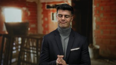 Arka planında tuğla duvarlar olan, gözlüklü ve balıkçı yaka giyen İspanyol kökenli ve kapalı alanda kendine güvenen bir ifade sergileyen bir inşaat alanında el çırpan genç adam..