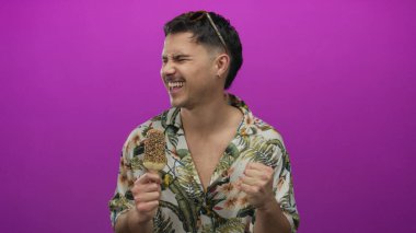 İspanyol asıllı genç adam pembe arka planda, çiçekli gömlekli ve modern tarzda neşeli yaz kaçışını yansıtan pembe elbisesiyle dondurma keyfine varıyor.