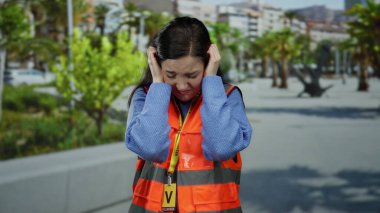 Turuncu yelekli kadın, arka planda yeşil ağaçlar olan kalabalık caddedeki gürültüden kulaklarını kapatıyor ve dışarıda rahatsızlığını dile getiriyor..
