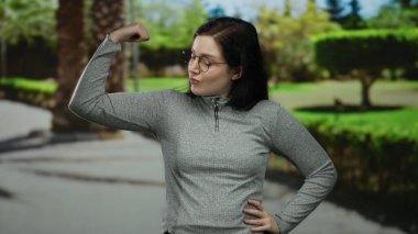 Parktaki kadın kollarını kavuşturarak gücünü gösteriyor. Çevresi yeşilliklerle çevrili. Açık havada güçlü ve eğlenceli bir an yakalıyor..