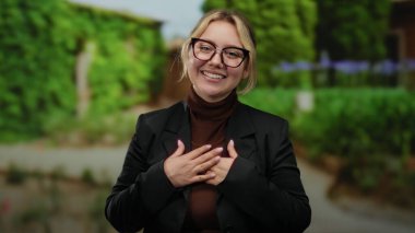 Açık hava parkı ortamında gülümseyen yeşil arka planda gözlük takan ve siyah ceketli bir kadın doğal bir ortamda pozitiflik ve rahatlama sergiliyor.