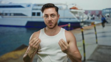 Sakallı genç bir adam, arka planda demirlemiş teknelerle dolu bir deniz kıyısındaki beyaz bir kolsuz bluzla rahat ve açık deniz atmosferi yaratıyor..