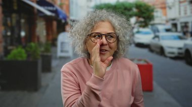 Şehir sokaklarında gülümseyen ve konuşan kır saçlı kadın, hareketli bir şehir ortamında olgun bir özgüvenin ve açık hava aktivitesinin özünü yakalıyor..