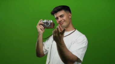İspanyol asıllı genç bir adam elinde bir kamera tutuyor ve canlı yeşil bir duvar arkaplanına karşı bir jest yapıyor, büyüleyici ve davetkar bir etkileşim..
