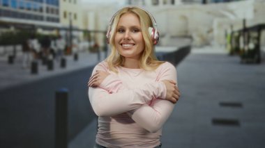 Kulaklık takan genç sarışın kadın şehir caddesinde durup sakin ve düşünceli bir şehir manzarası yaratırken kendini kucaklıyor..