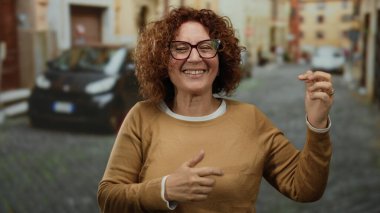 Güneşli bir caddede gitar çalan, gülümseyen ve el kol hareketi yapan bir kadın. Arka planda bulanık arabalar ve tarihi binalar var. Neşeli şehir atmosferini yakalıyor..