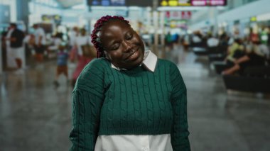 Havaalanı terminalinde oturan yeşil kazaklı kadın, mutlu bir ifadeyle, kalabalık gezginlerle çevrili, kapalı bir havaalanı ortamında..