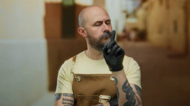 Sakallı, dövmeli, önlüklü ve eldivenli bir adam açık hava ortamını işaret ediyor. Gözleri kapalı, şehir sokaklarında odaklanma ve kararlılık gösteriyor..