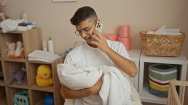 Bebek odasında bebeğini kucağında tutan yakışıklı bir adam telefonda konuşurken, oyuncaklarla ve bebek malzemeleriyle çevrili, rahat ve şefkatli bir ortam sergiliyor..
