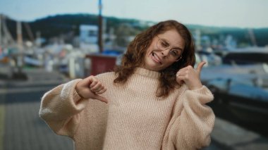 Deniz kıyısındaki bir kadın karışık jestler yapıyor, gözlük ve süveter takıyor, etrafı tekne ve manzarayla çevrili..