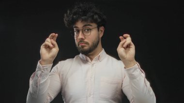 Sakallı ve gözlüklü genç bir adam siyah bir arka plana, bir stüdyo ortamında yüz ifadesine ve jestlere karşı tırnak işareti yapıyor..