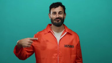 Turuncu hapishane tulumu giymiş sakallı İspanyol bir adam yeşil bir arkaplanda sırıtışıyla kendini işaret ediyor..