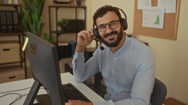 Modern ofis ortamında bilgisayar üzerinde çalışan sakallı İspanyol bir adam. Etrafı bitki ve ofis dekorlarıyla çevrili bir kulaklık takıyor. Çağdaş bir çalışma ortamı yaratıyor..