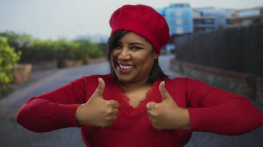 Şehir caddesinde gülümseyen, kırmızı bereli ve kazaklı, başparmak kaldıran, açık şehir ortamında kendine güveni somutlaştıran, genç, iri yarı Latin bir kadın..