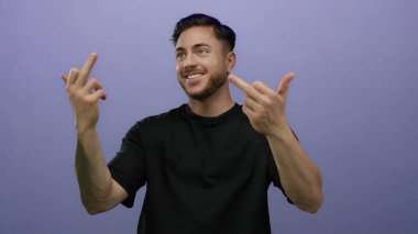 Sakallı genç adam gülümserken orta parmaklarıyla jestler yapıyor, izole edilmiş mor arka plana karşı duruyor..