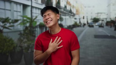 Şehrin sokaklarında neşeli bir şekilde gülen genç bir adam kırmızı tişört giyip açık havada mutluluk ve enerjiyi somutlaştırıyor.