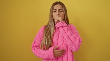 Soğuk ya da bronşitten rahatsız olan, ayakta duran ve izole edilmiş sarı arka plan üzerinde öksüren genç sarışın kadın sağlık sorunlarını vurguluyor.